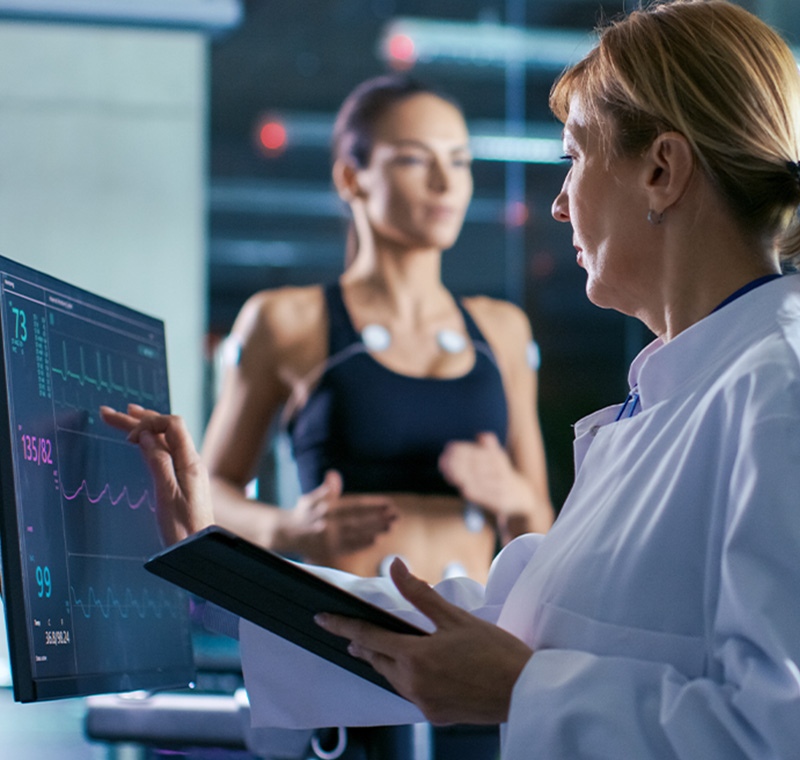 A cardio phlebotomy technician in medical scrubs gently draws blood from a patient’s arm while monitoring heart rate equipment, demonstrating skills in both phlebotomy and cardiovascular testing.
