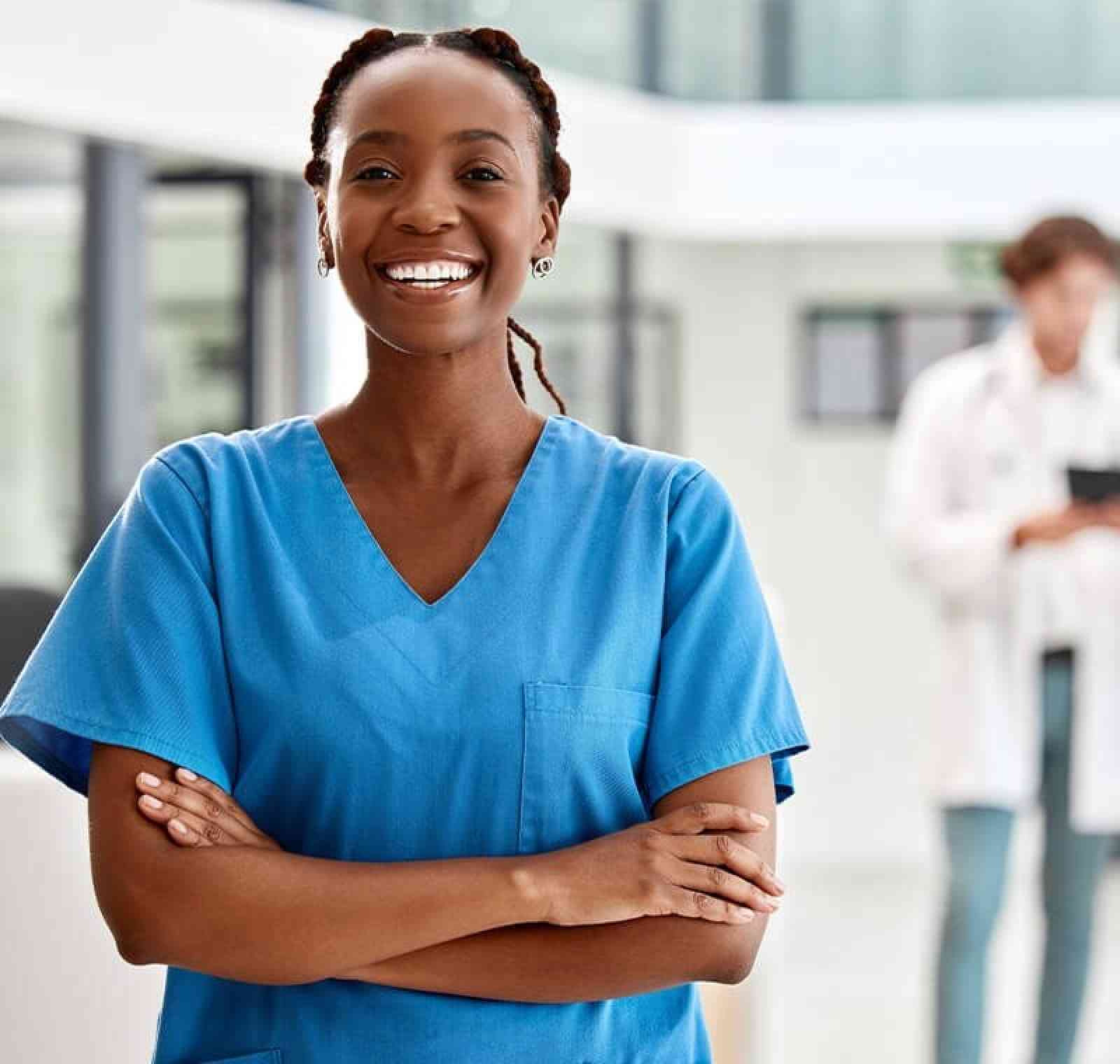 A smiling medical assistant in blue scrubs stands in a modern clinical setting with arms crossed, confidently representing a medical-assistant role. A doctor and other clinical details blur softly in the background.