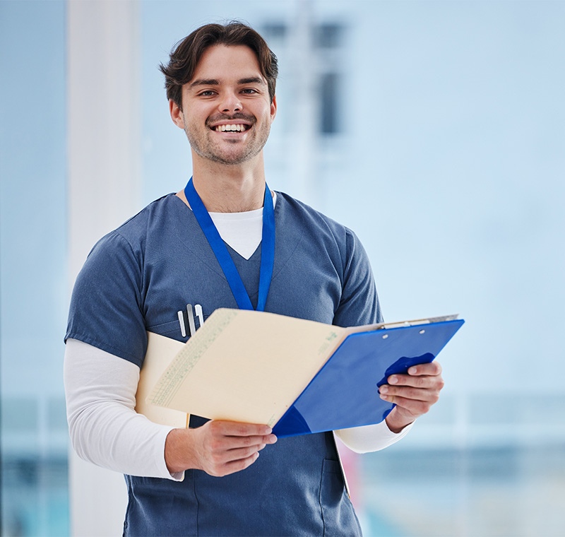 A medical front office assistant greets a patient at the reception desk, entering information into an electronic health record system in a bright, welcoming clinic environment.